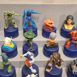 May include: A collection of colorful Gundam robot figurines displayed on blue bottle caps. The figures vary in design and color, including blue, green, orange, and white, and are arranged on a gray shelf.