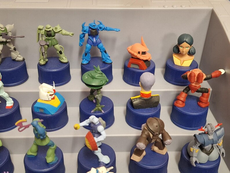 May include: A collection of colorful Gundam robot figurines displayed on blue bottle caps. The figures vary in design and color, including blue, green, orange, and white, and are arranged on a gray shelf.