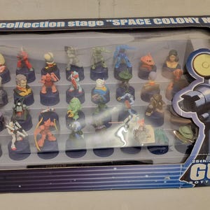 May include: A collection of colorful miniature figures displayed in a clear plastic case. The figures are on small, dark blue bases. The packaging reads "The collection stage SPACE COLONY MODEL" and "25th Anniversary GUNDAM Bottle Cap Collection".