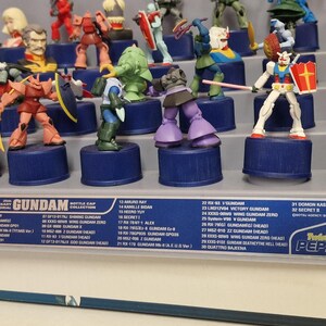 May include: A collection of Gundam bottle cap figures displayed on a shelf. The figures are of various colors and designs, mounted on blue bottle caps. The display includes a list of the figures.