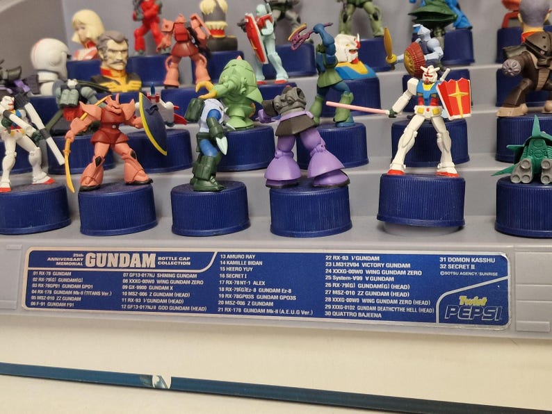 May include: A collection of Gundam bottle cap figures displayed on a shelf. The figures are of various colors and designs, mounted on blue bottle caps. The display includes a list of the figures.