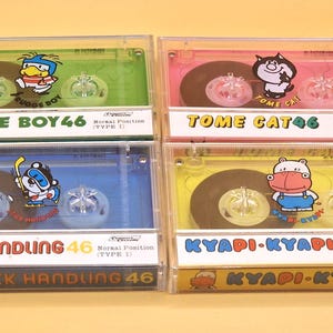 May include: Four clear plastic cassette cases containing colorful audio tapes in green, pink, blue, and yellow. Each tape features a cartoon character and text labels including Rugge Boy 46, Tome Cat 46, Stick Handling 46, and Kyapi-Kyapi 46, all marked Type I.