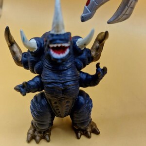 Puede incluir: Una figura de monstruo azul oscuro con m&uacute;ltiples cuernos, brazos y piernas. La figura tiene una amplia sonrisa dentuda y detalles dorados en los brazos y los pies. Otra garra de monstruo gris es visible en el fondo.