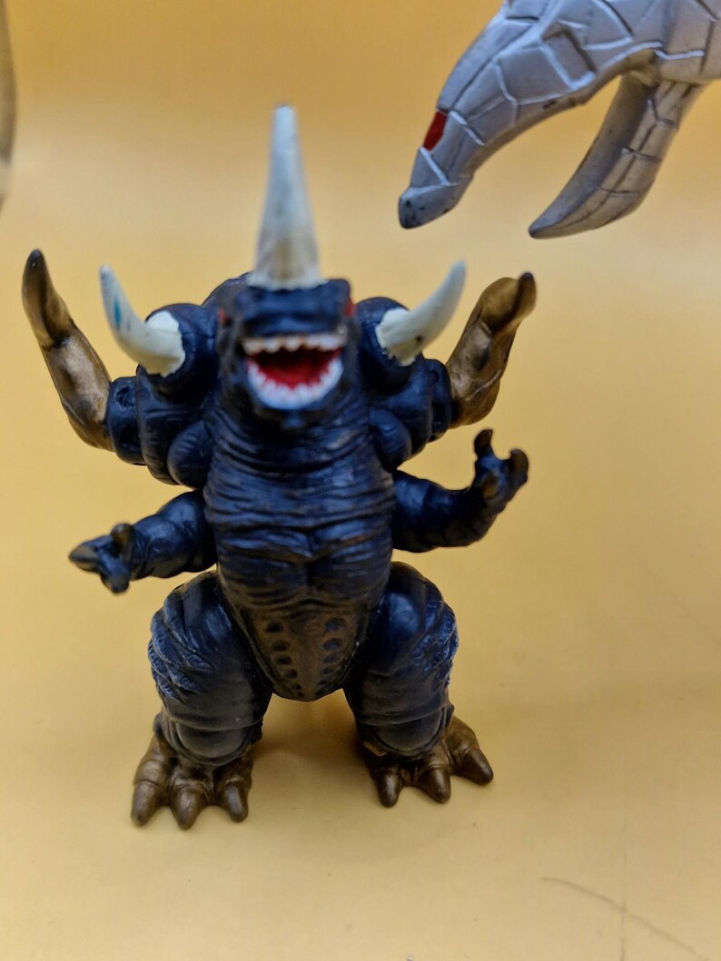 Puede incluir: Una figura de monstruo azul oscuro con m&uacute;ltiples cuernos, brazos y piernas. La figura tiene una amplia sonrisa dentuda y detalles dorados en los brazos y los pies. Otra garra de monstruo gris es visible en el fondo.