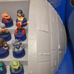 May include: A collection of colorful miniature figurines, each mounted on a blue cylindrical base. The figurines depict various characters, with details in red, orange, blue, and green. A large, gray, spherical structure is visible on the right side of the image.