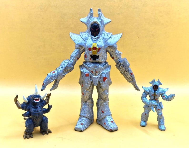 Puede incluir: Tres figuras de acci&oacute;n de juguete. Una gran figura de robot plateado con detalles rojos se encuentra entre un robot plateado m&aacute;s peque&ntilde;o y un monstruo azul con cuernos y m&uacute;ltiples brazos. Las figuras est&aacute;n sobre un fondo amarillo.