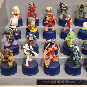 May include: A collection of Gundam bottle cap figures, featuring various characters in dynamic poses. The figures are brightly colored, with details like weapons and armor. Each figure is mounted on a blue bottle cap.