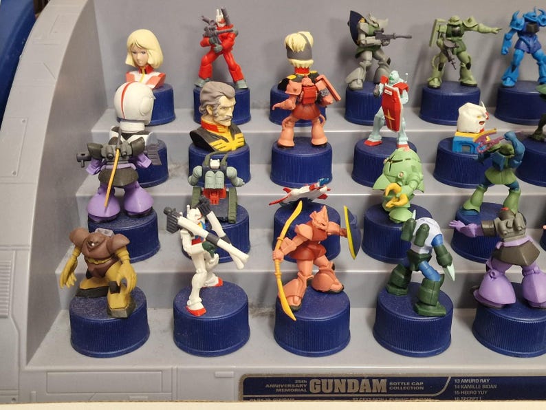 May include: A collection of Gundam bottle cap figures, featuring various characters in dynamic poses. The figures are brightly colored, with details like weapons and armor. Each figure is mounted on a blue bottle cap.