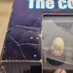 May include: A partially visible, damaged box with the text "The col..." in white letters on a blue background. The box contains small figurines, some with red and white details, visible through a clear plastic window.