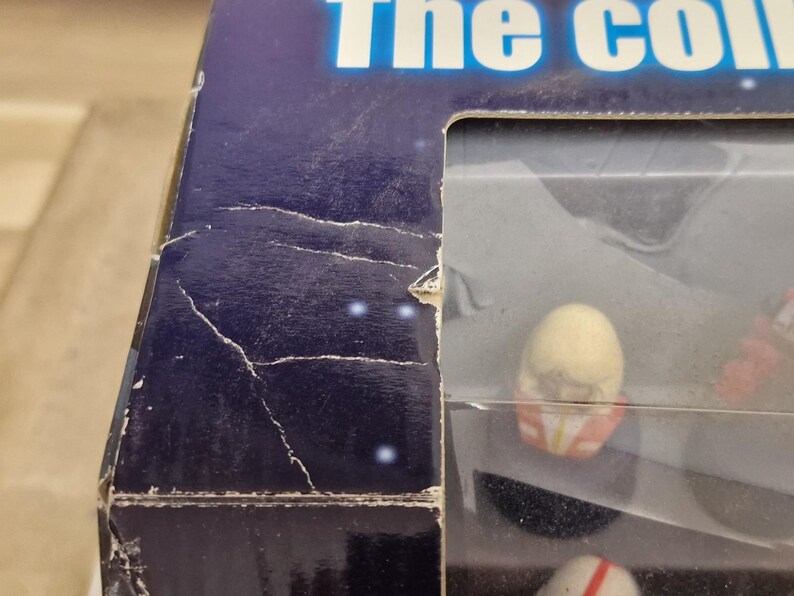 May include: A partially visible, damaged box with the text "The col..." in white letters on a blue background. The box contains small figurines, some with red and white details, visible through a clear plastic window.