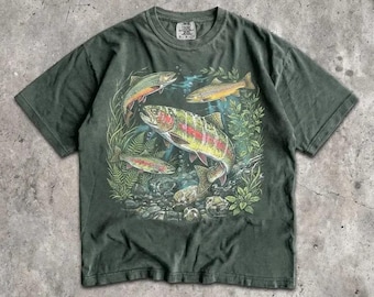 May include: A dark olive green short-sleeve t-shirt featuring a graphic print of three trout swimming among aquatic plants and rocks. The shirt has a crew neckline and a Comfort Colors brand label printed on the inside of the collar area.