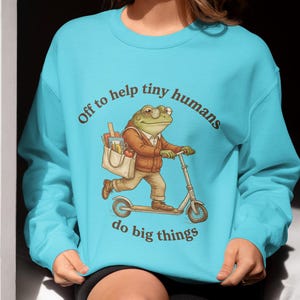 Peut inclure: Un sweat-shirt turquoise avec un dessin animé d'une grenouille sur un scooter, portant des lunettes et une veste, avec un sac. Le texte dit "Off to help tiny humans do big things."
