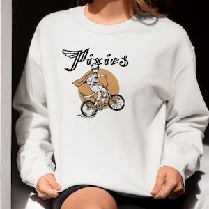 May include: White sweatshirt featuring a graphic of a person riding a bicycle with the word "Pixies" in black lettering.