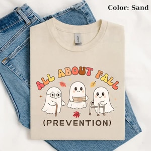 May include: A sand-colored t-shirt featuring a graphic of three white ghosts. One ghost uses a cane, one wears a belt, and one uses a walker. The text reads ALL ABOUT FALL (PREVENTION) in colorful letters, accented by small red and yellow autumn leaves.