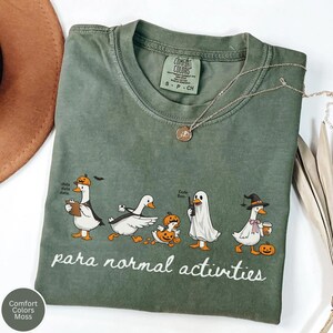 May include: A moss green t-shirt featuring a graphic of five white ducks in various Halloween costumes, including a pumpkin, ghost, and witch hat. The text "para normal activities" is printed below the ducks in a white script font.