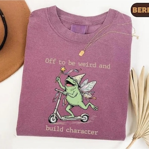 Könnte beinhalten: Ein beerenfarbenes T-Shirt mit einer Grafik eines grünen Frosches mit Feenflügeln und spitzem Hut auf einem Roller, der einen Zauberstab mit Stern hält. Der Text lautet, Off to be weird and build character. Eine goldene Halskette liegt am Kragen.