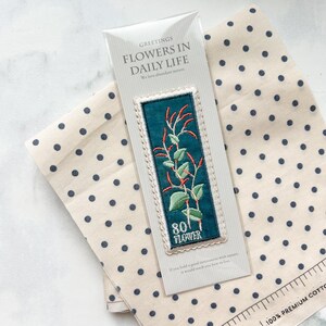 May include: A bookmark with an embroidered floral design, featuring red and green accents on a teal background. The bookmark is packaged with a card that reads "FLOWERS IN DAILY LIFE". The background includes a beige fabric with navy blue polka dots.