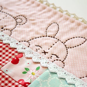 May include: A pink fabric with white polka dots and a white crocheted trim. The fabric has embroidered designs of a bear, a bunny, and a butterfly.