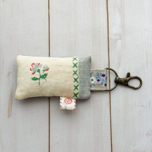 May include: A small, rectangular keychain with a floral design. The cream-colored fabric features a red and green flower embroidery. The keychain has a gray border with green cross-stitch details and a small tag with text. A bronze-colored clasp is attached.