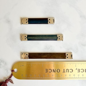 May include: Three rectangular wooden pieces with metal corner accents. The pieces are various shades of brown and blue-green. A brass ruler with the text "NICE, CUT ONCE" is next to the pieces, measuring in inches.