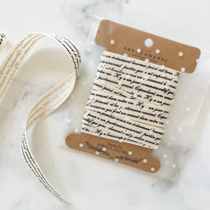 Peut inclure: Un rouleau de ruban crème avec du texte en noir, emballé dans un sac en plastique transparent avec une étiquette marron. Le ruban est déroulé, montrant le motif du texte. L'étiquette indique "nan Company" et "SPECIAL DELIVE".