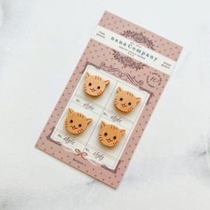May include: Four wooden cat-shaped buttons with etched details, arranged on a pink card. The buttons have a light brown color with etched facial features. The card has the text "nana Company" and "Pretty Little Things".