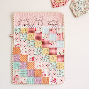 May include: A pink and white patchwork baby blanket with a crocheted border. The blanket features a variety of floral and geometric patterns. The top of the blanket has a pink fabric with embroidered animal faces.
