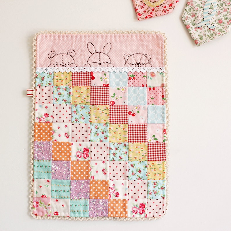 May include: A pink and white patchwork baby blanket with a crocheted border. The blanket features a variety of floral and geometric patterns. The top of the blanket has a pink fabric with embroidered animal faces.