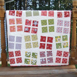 May include: A white quilt with a patchwork design featuring red, green, and blue squares with various patterns. The quilt is hanging on a wooden railing.