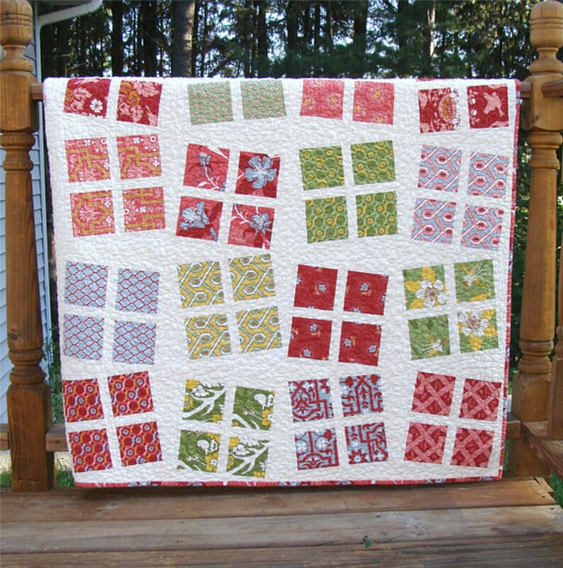 May include: A white quilt with a patchwork design featuring red, green, and blue squares with various patterns. The quilt is hanging on a wooden railing.