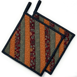 Puede incluir: Dos agarraderas de olla acolchadas con un patrón a rayas en tonos naranja, verde y rojo. Las agarraderas tienen un ribete azul oscuro y un lazo para colgar.