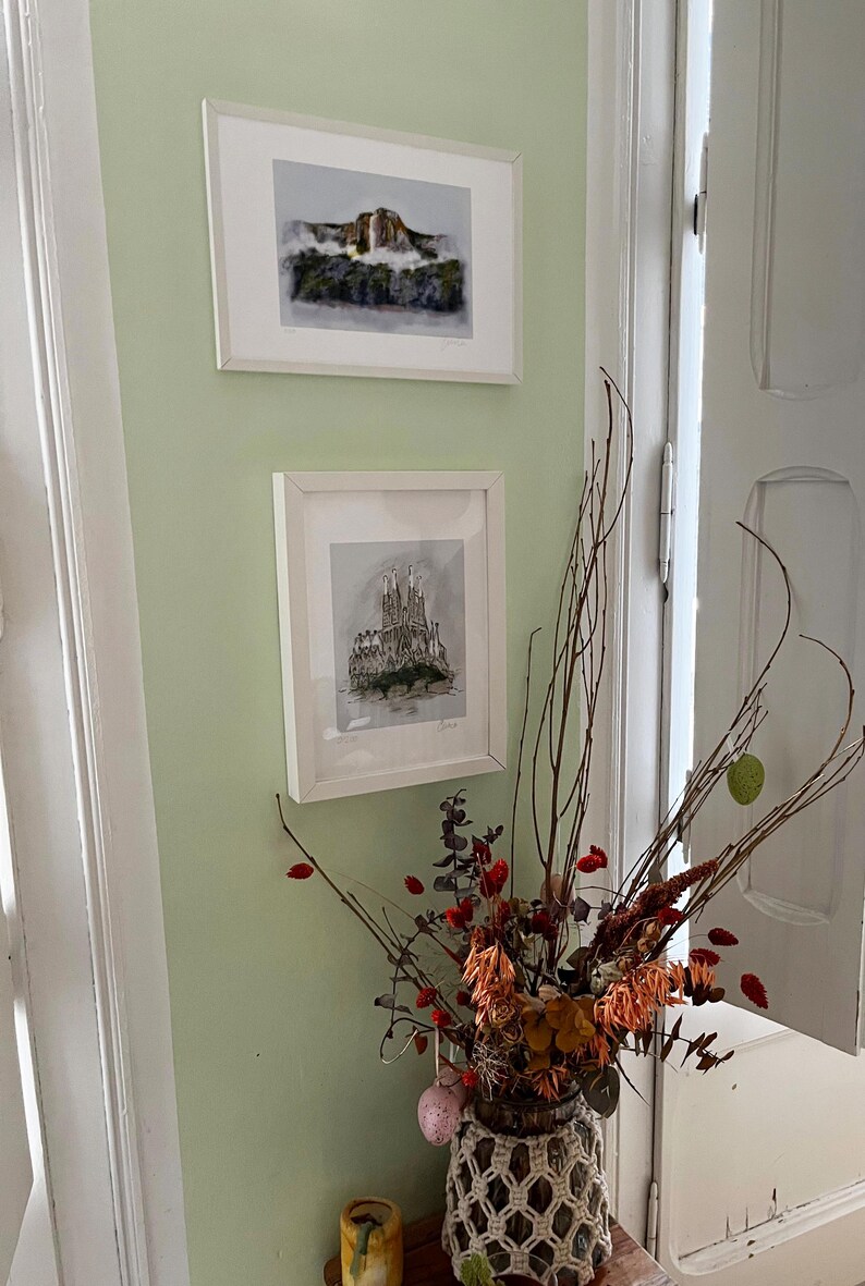 May include: Two framed black and white drawings of architectural structures hang on a pale green wall. The top drawing depicts a waterfall, while the bottom drawing shows a building with a pointed roof. A vase with dried flowers sits on a table in the foreground.