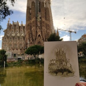 May include: A postcard with a black and white sketch of the Sagrada Familia church in Barcelona, Spain. The postcard is held in front of the actual Sagrada Familia church.
