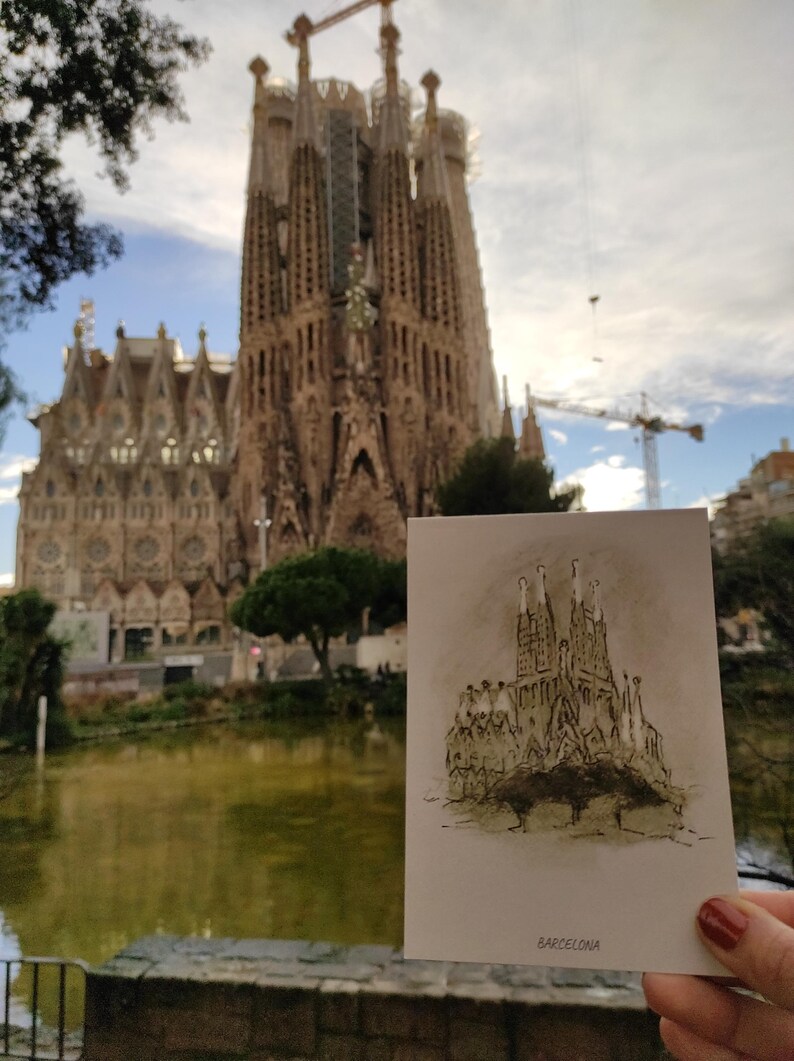 May include: A postcard with a black and white sketch of the Sagrada Familia church in Barcelona, Spain. The postcard is held in front of the actual Sagrada Familia church.