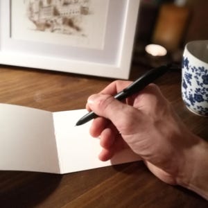 May include: A person's hand holding a black pen over a blank white card. The card is folded in half and is sitting on a wooden table.