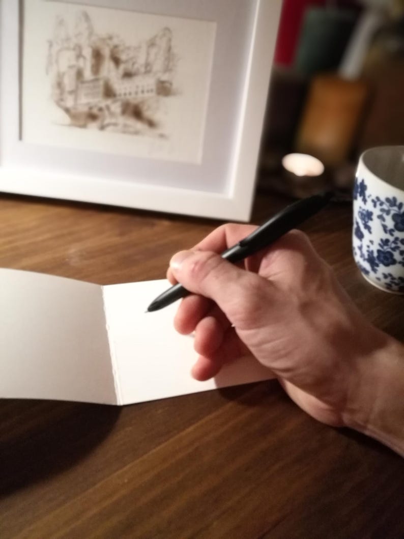 May include: A person's hand holding a black pen over a blank white card. The card is folded in half and is sitting on a wooden table.