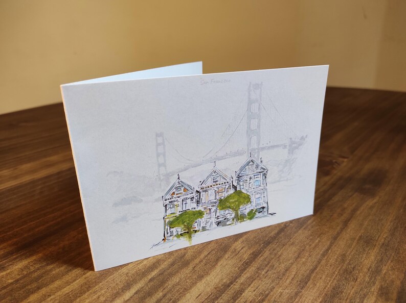 May include: A watercolor painting of three colorful houses with green trees in front of them. The houses are painted in a whimsical style and the background is a light blue with a faint outline of the Golden Gate Bridge.