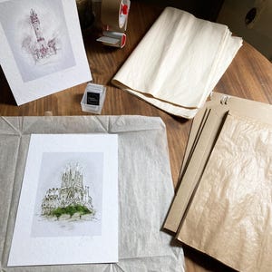 May include: A white mount with a black and white ink drawing of a building on top of it. The mount is on top of a brown paper bag. There is a roll of brown paper and a roll of white paper in the background.