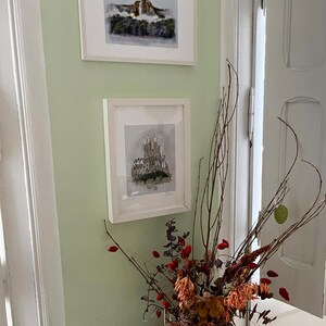 May include: Two framed black and white drawings of architectural structures hang on a pale green wall. The top drawing depicts a waterfall, while the bottom drawing shows a building with a pointed roof. A vase with dried flowers sits on a table in the foreground.