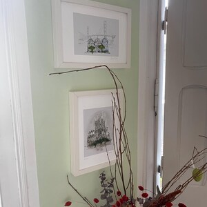 May include: Two framed black and white illustrations of cityscapes hanging on a pale green wall. The top illustration shows a bridge with buildings in the foreground. The bottom illustration shows a tall building with a pointed roof.