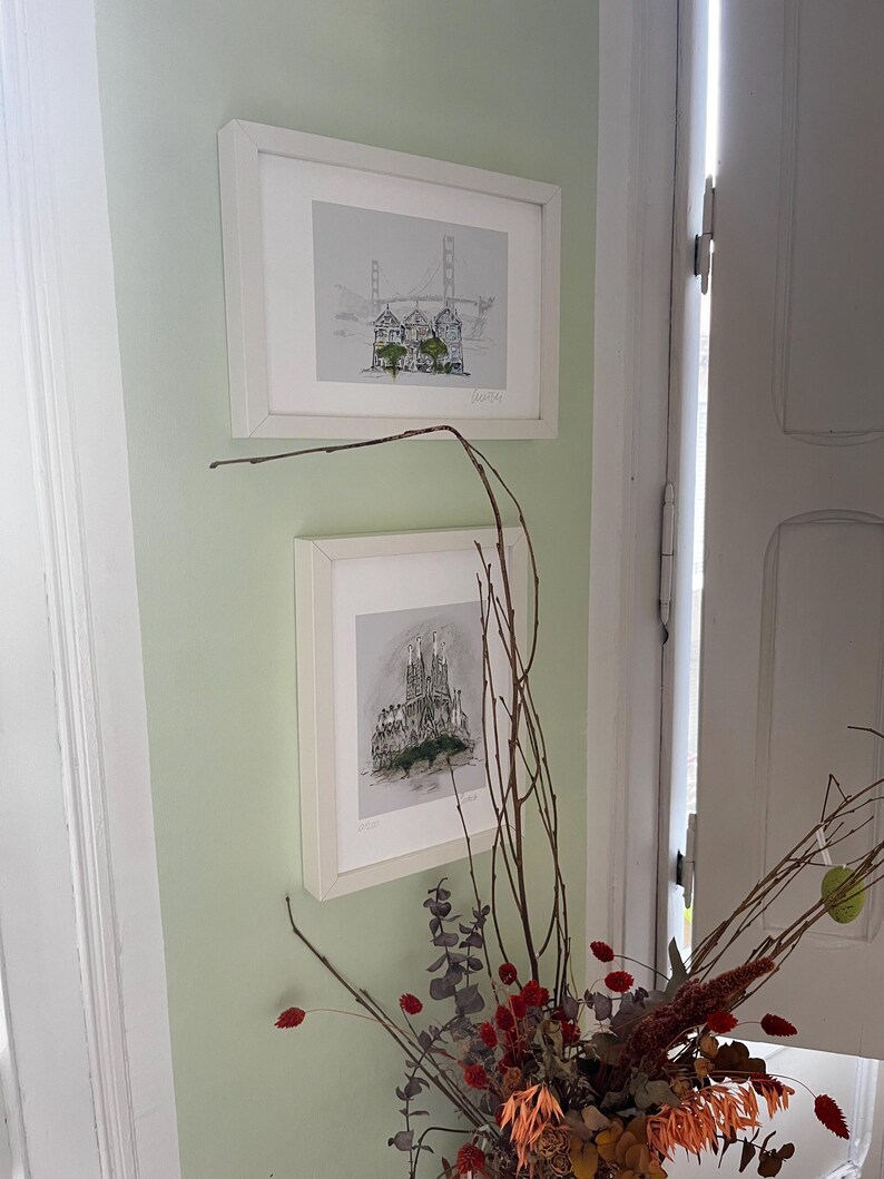 May include: Two framed black and white illustrations of cityscapes hanging on a pale green wall. The top illustration shows a bridge with buildings in the foreground. The bottom illustration shows a tall building with a pointed roof.