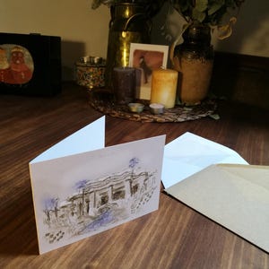 May include: A blank greeting card with a watercolor illustration of a building with a blue sky and trees. The card is on a wooden table with a brown envelope next to it.