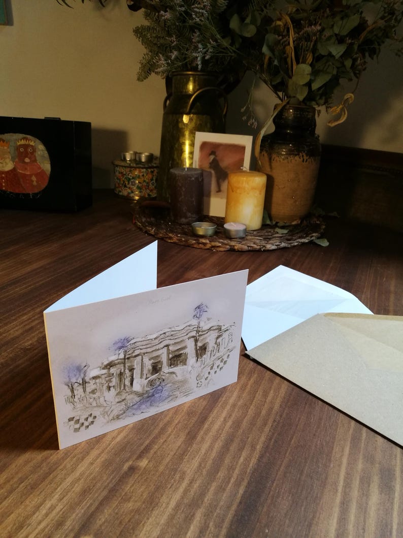 May include: A blank greeting card with a watercolor illustration of a building with a blue sky and trees. The card is on a wooden table with a brown envelope next to it.