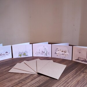 May include: Six blank greeting cards with different cityscapes drawn on them. The cards are arranged in a row on a wooden table. There are also six blank brown envelopes on the table.