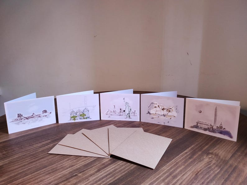 May include: Six blank greeting cards with different cityscapes drawn on them. The cards are arranged in a row on a wooden table. There are also six blank brown envelopes on the table.