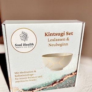 Könnte beinhalten: Eine weiße Schachtel mit den Wörtern "Kintsugi Set" und "Soul Health" aufgedruckt. Die Schachtel zeigt eine Illustration einer weißen Schale mit goldfarbenen Rissen. Der Text "Moments for inner Balance" ist ebenfalls sichtbar.