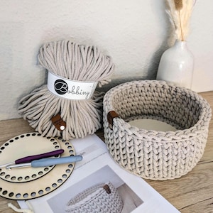 Könnte beinhalten: Ein beige gehäkelter Korb mit Lederakzent steht neben einem Knäuel beiger Wolle mit der Aufschrift "Bobbiny". Zwei Häkelnadeln und Holzsockel sind ebenfalls zu sehen, zusammen mit einer gedruckten Anleitung. Die Artikel deuten auf ein DIY-Bastelprojekt hin.