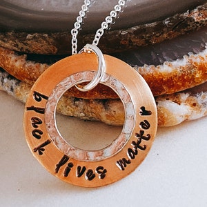 May include: A copper-colored pendant necklace with the words "black lives matter" stamped around the outer edge. The pendant has a circular design with a textured, light-colored inner ring and a silver chain.