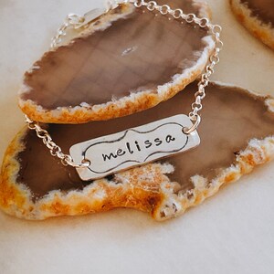 May include: A silver bracelet with a rectangular pendant. The pendant has a decorative border and the name "melissa" is handwritten in black. The bracelet is displayed on a brown and orange agate slice.