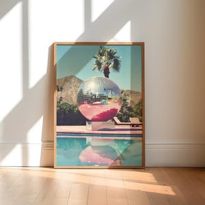 May include: A large silver disco ball sits in a swimming pool with a palm tree in the background. The pool is surrounded by a pink tiled deck.
